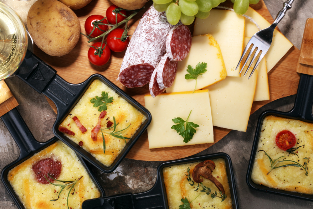 Raclette-Pfännchen mit Käse neben einer Holzplatte mit Salami, Käse und Gemüse