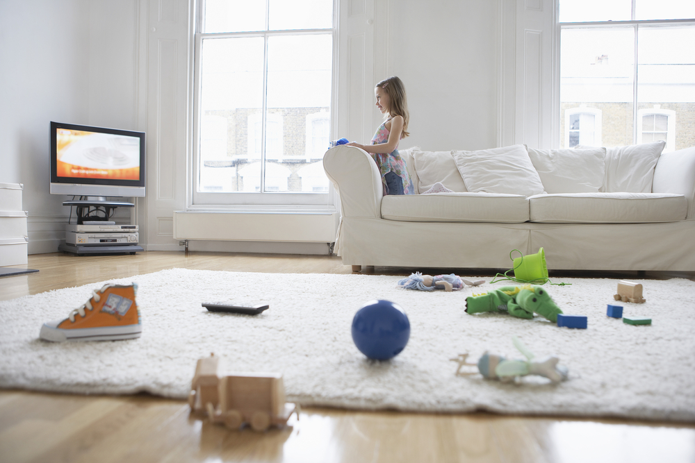Mädchen auf weißer Couch schaut TV, auf dem weißen Teppich liegt diverses Spielzeug