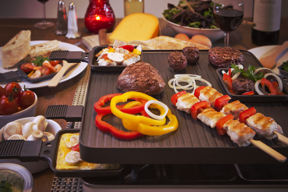 Raclette-Grill mit Fleisch und Gemüse belegt, dahinter ein Stück Käse