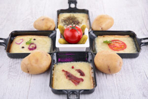 Vier Raclette Pfännchen, daneben Kartofflen mit Schale und in der Mitte eine Tomate