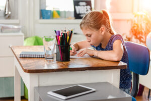 Junges Mädchen im blauen Shirt an einem Holztisch beim Erledigen der Hausaufgaben