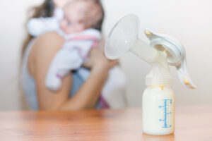 Im Vordergrund eine manuelle Milchpumpe, im Hintergrund verschwommen eine Mutter mit Baby im Arm