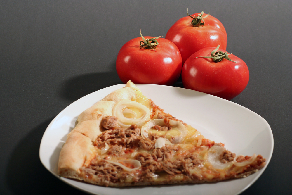 Eine Pizzaecke auf weißem Teller, dahinter drei Tomaten