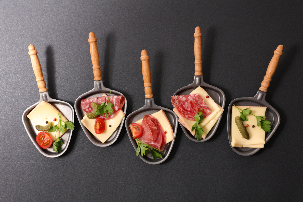 Fünf mit Schinken und Käse gefüllte Raclette-Pfännchen auf schwarzem Hintergrund